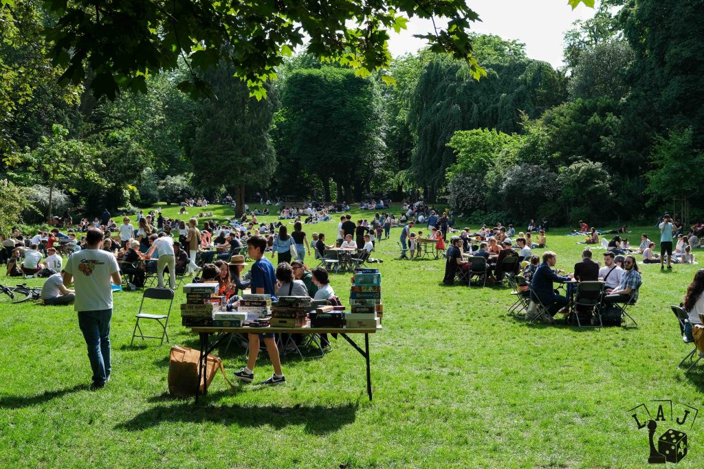 Photo de l’édition 2025 de Lille aux Jeux au Jardin Vauban. On y voit de nombreuses personnes rassemblées dans un parc verdoyant, assises autour de tables ou sur l’herbe pour jouer à des jeux de société. 
